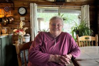 A woman in a maroon longsleeve shirt and glasses, sits at a table with her hands folded. A portable oxygen machine is helping her breathe