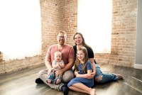 A mother and father sit on the floor in a well-lit room with their two young children in their laps for a studio portrait.
