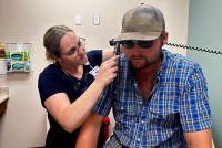 A female nurse practitioner, in wire frame glasses, inspects the ear of a man in a cap, sunglasses, and a check shirt.