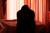 A person sitting on a bed, their head in their hands, backlit by a window