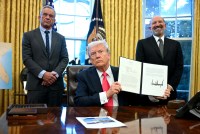 A photo of President Trump showing a signed executive order to photographers in the Oval Office of the White House. He is flanked by Robert F. Kennedy Jr. and Howard Lutnick.
