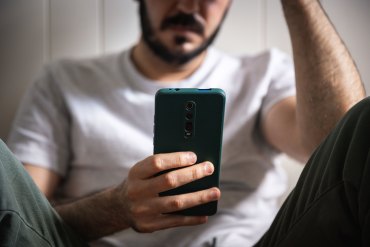 A man with a beard and wearing jeans and a t-shirt sits on the floor and looks at a phone in his hand.