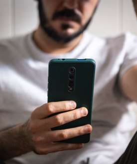 A man with a beard and wearing jeans and a t-shirt sits on the floor and looks at a phone in his hand.