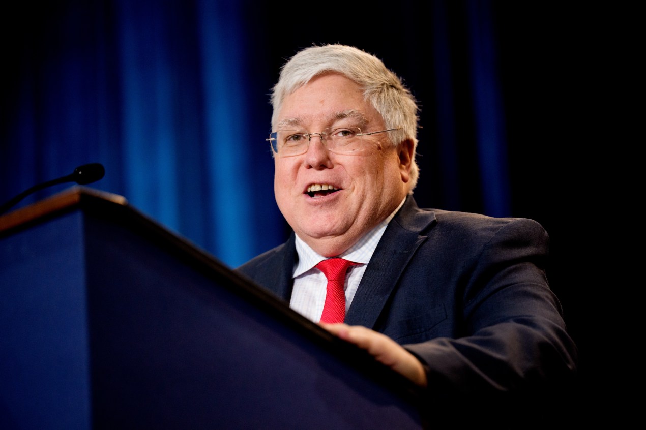 A man with grey hair, glasses, a red tie and a dark suit stands behind a podium