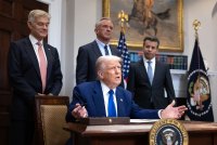 A photo of President Trump seated at a desk, gesturing with his hands, holding a magic marker. Three men stand behind him; they are heads of federal health agencies.
