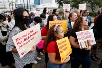 A group of people stand together and hold signs that read "Nurses Care For ALL People," "Our Patients Rights Have No Borders," or "Nurses Care. No Exceptions." and the logo and name of National Nurses United on them.