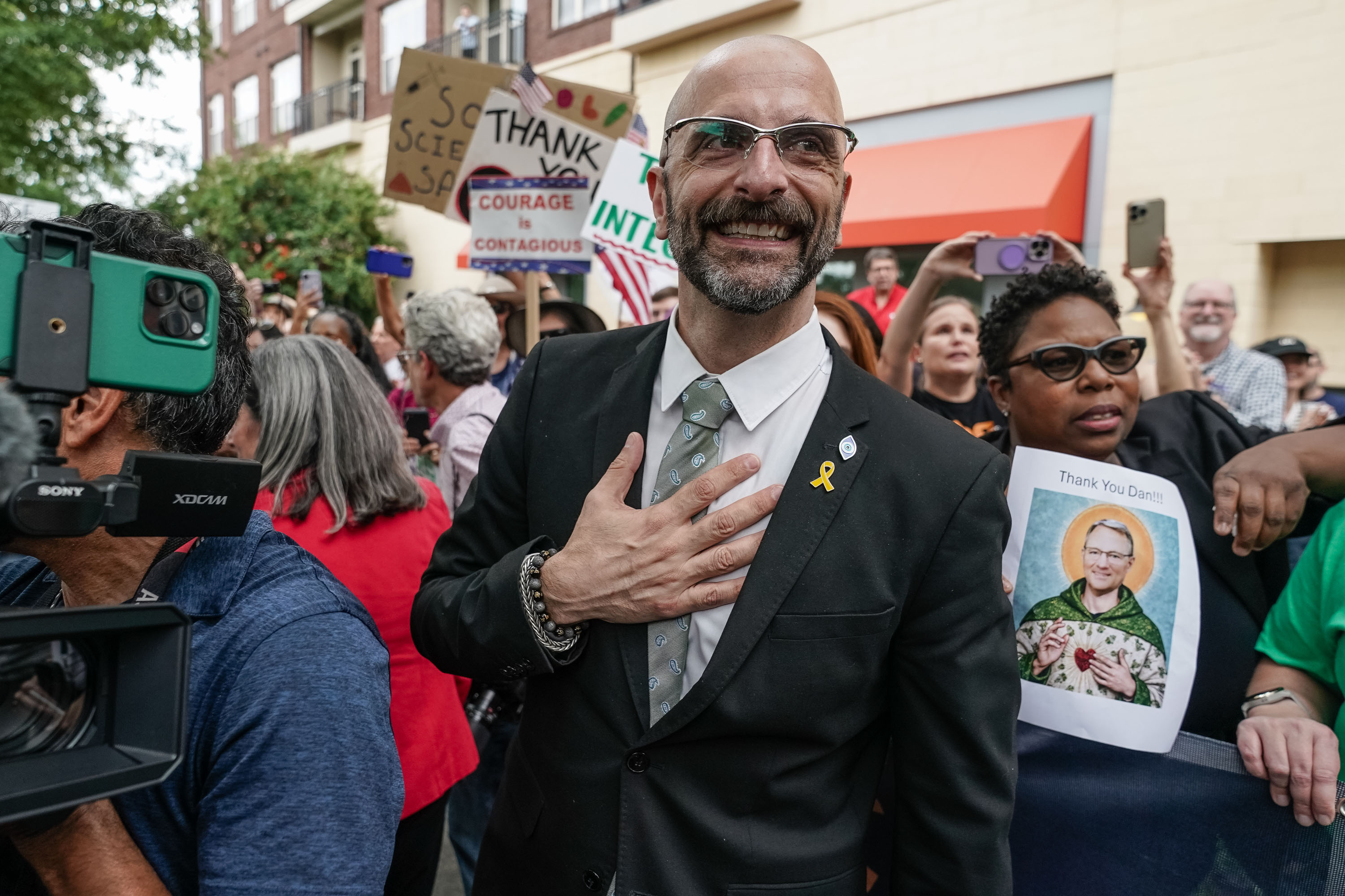 A photo of a man wearing a suit and tie as he stands in a crowd. He puts his left hand to his chest and smiles.