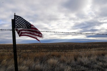 An American flag blows in the wind next to a barbed-wire fence in front of a landscape of grasslands