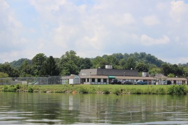 In a photo taken from a craft on a river, a single-story building building sits near the water. There is a fenced-in playground, a shed, a gazebo, and a small parking lot surrounding the building. Trees can be seen in the distance behind the building and its grounds.