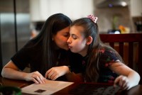 A woman with dark brown hair sits at a table and touches her face to the face of a girl wearing a pink bow in her hair
