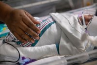A close-up photo of a hand on a swaddled baby in an ICU bed.