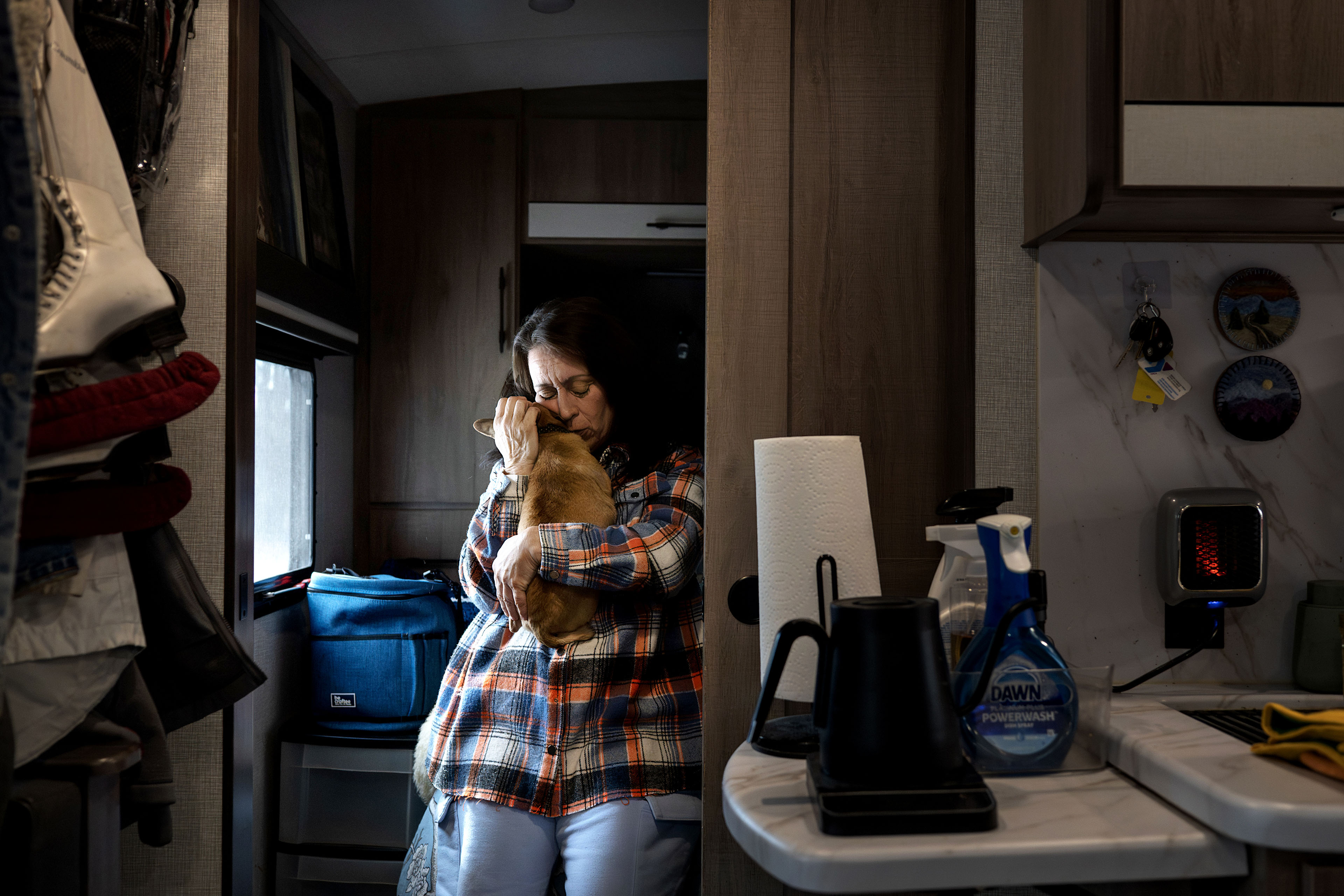 A woman stands in her kitchen while holding her small dog tenderly to her chest, kissing its head.