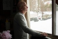 A side-view of a man standing at a window and looking out. His right had rests on the window sill.