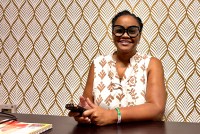 A woman wearing black-rimmed glasses and a white-and-brown patterned top sits at a table holding a cell phone while posing for a portrait