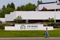 A sign in front of a building reads "Covered California." Two people walk along the sidewalk in front of the sign.