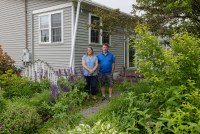 A husband and wife stand outside their home, surrounded by lush green plants and tall purple flowers.