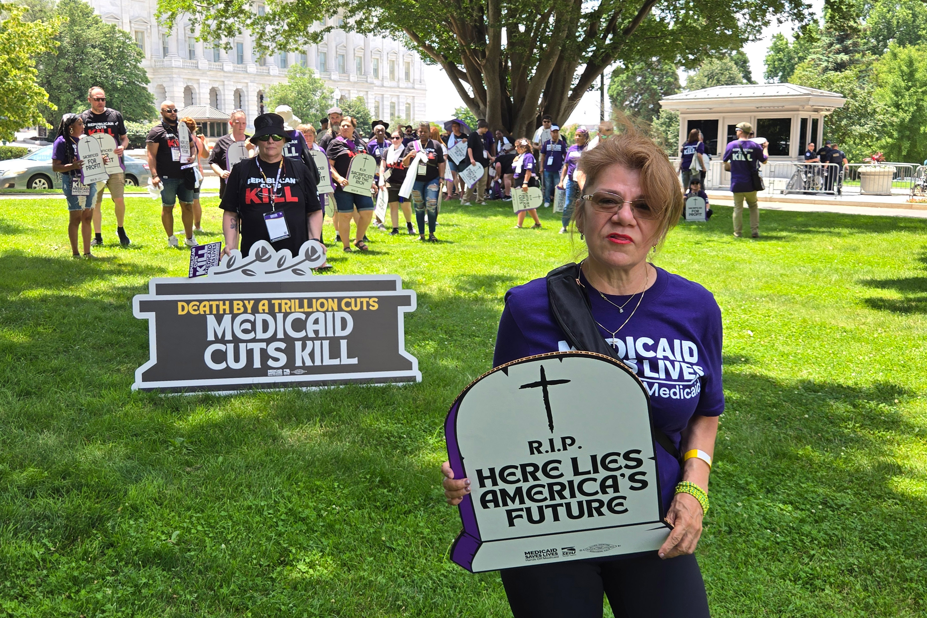 A photo of a woman standing for a portrait outside. She holds a protest sign shaped like a tombstone reading, "Here lies America's future." Other protesters are seen in the background.