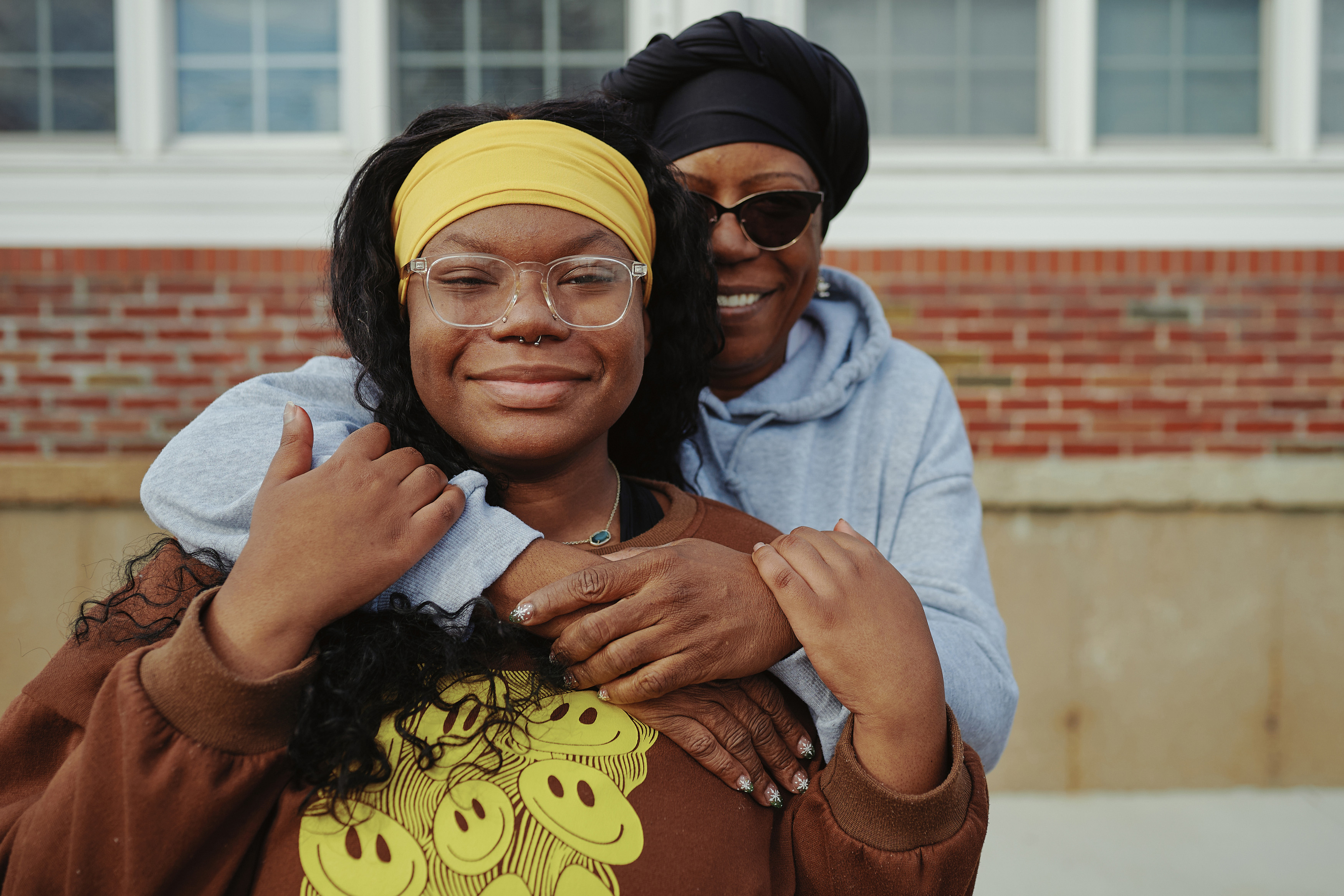 A woman wearing sunglasses and a black hat stands behind and puts her arms around a woman wearing glasses and a yellow headband as both smile for a portrait