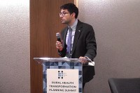 A photo of a man standing at a podium and speaking into a microphone.