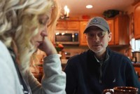 A husband sits at the kitchen table. His wife is seen close on the left of the frame. She is blurred slightly as the camera is focused on the husband.