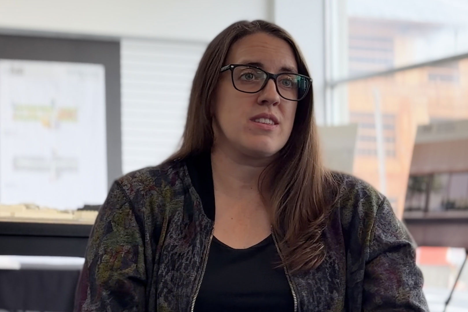 A woman with long dark hair and glasses sits and speaks to someone just off-camera. She wears a dark jacket and shirt.